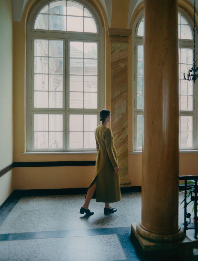 A woman in a long coat walking in an interior hallway with large arched windows and marble pillars, bathed in soft, diffused light.