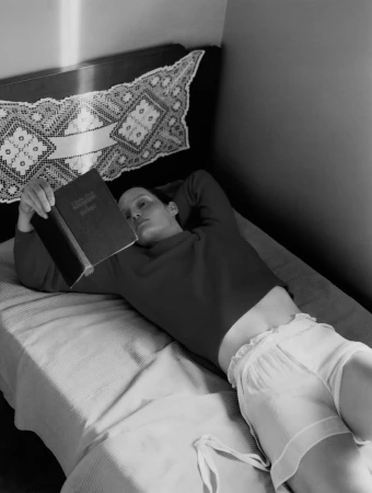 Model Vivien Solari lying on a bed holding an open book, wearing a dark crewneck sweater and white ruffled shorts in black and white.