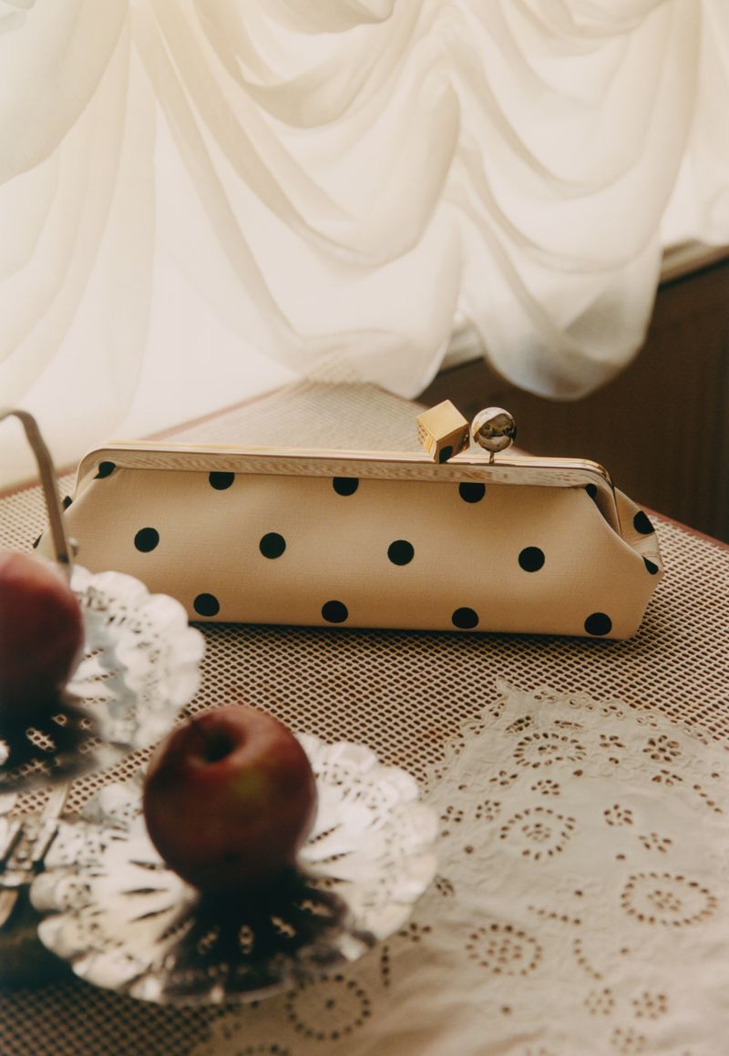 Close-up shot of a polka dot clutch bag on a decorative table next to two apples, with soft white curtains in the background.