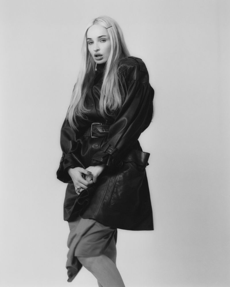 Kim Petras wearing a belted black leather trench coat posing against a studio seamless background in a black and white editorial shoot.