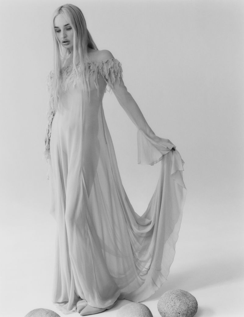 Kim Petras standing in a flowing sheer off-the-shoulder gown against a seamless studio backdrop with stones on the floor.