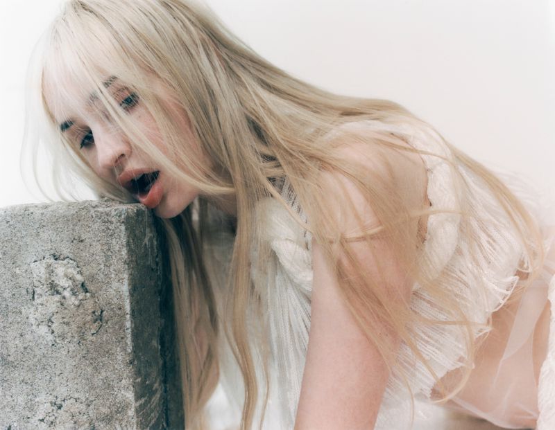 Kim Petras leaning her chin against a textured concrete block wearing a ruffled white top against a clean white backdrop.