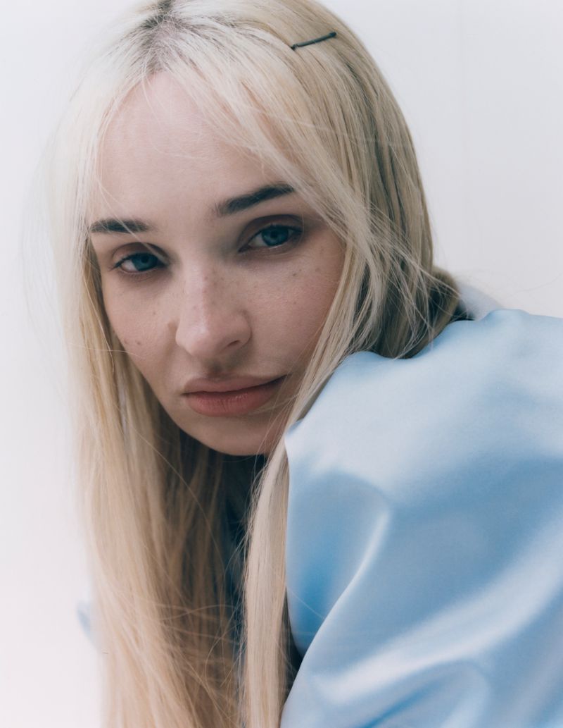 Close-up portrait of Kim Petras with blonde hair and bangs wearing a light blue satin garment against a white background.