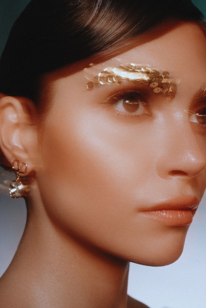 Close-up beauty portrait of a woman wearing gold sequin eyebrow makeup and a crystal drop earring.