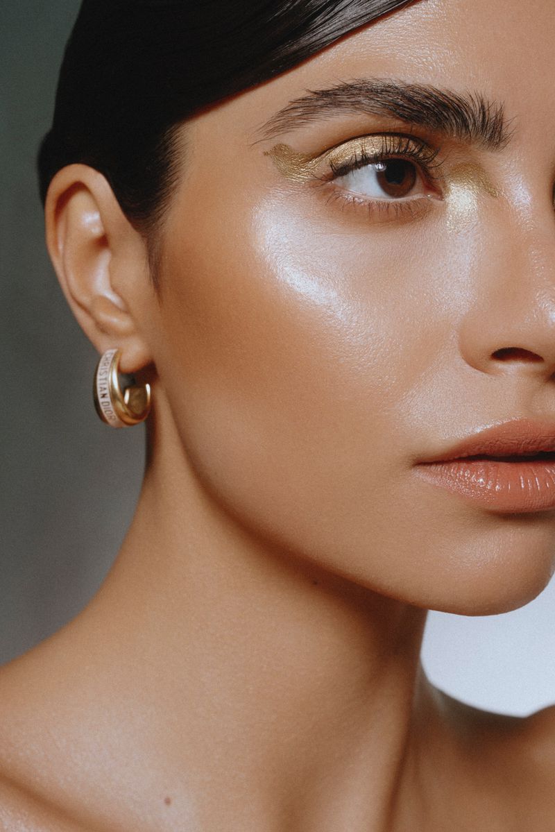 Close-up portrait of a woman with metallic gold eye makeup and hoop earrings against a neutral studio background