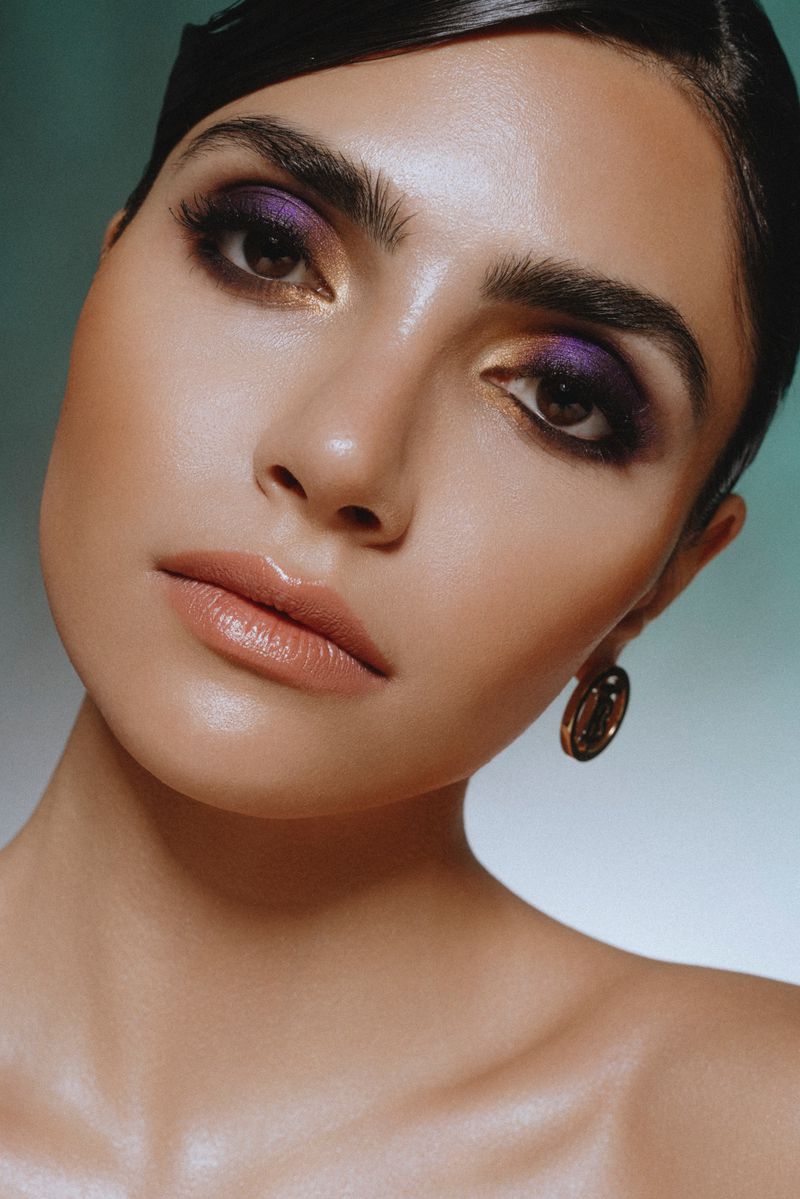 Close-up beauty portrait of a woman featuring purple and gold eyeshadow with glossy nude lips against a teal gradient background.