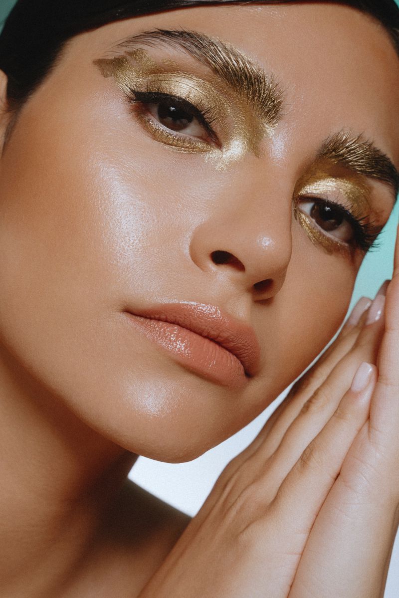 Close-up beauty portrait of a woman with shimmering gold eye makeup and painted gold eyebrows posing with hands near her face.