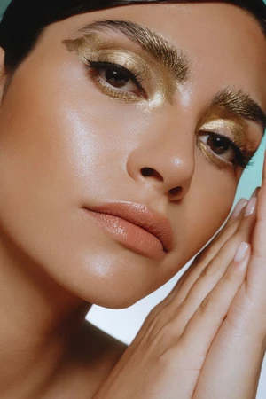 Close-up beauty portrait of a woman with shimmering gold eye makeup and painted gold eyebrows posing with hands near her face.