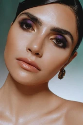 Close-up beauty portrait of a woman featuring purple and gold eyeshadow with glossy nude lips against a teal gradient background.
