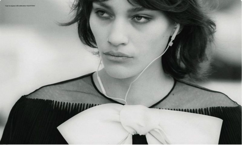 Close-up black and white portrait of a woman wearing wired earphones and a black top with an oversized white bow.