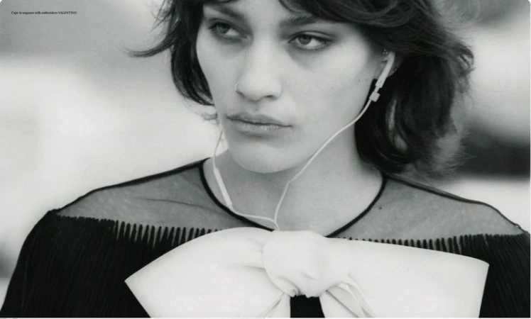 Close-up black and white portrait of a woman wearing wired earphones and a black top with an oversized white bow.