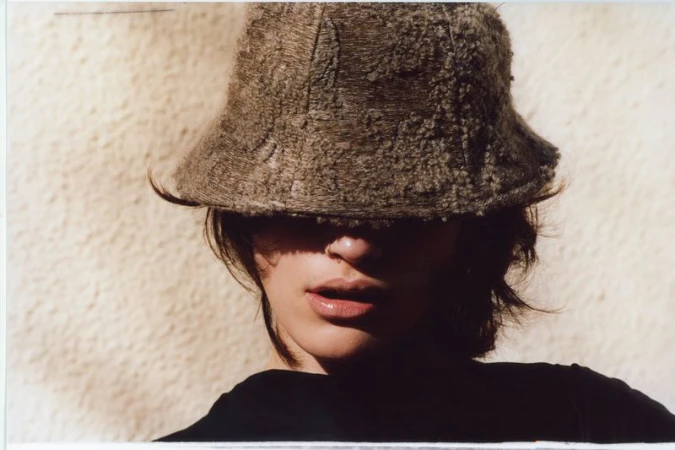 Close-up portrait of a model wearing a textured brown bucket hat pulled over their eyes against a sunlit neutral textured wall.