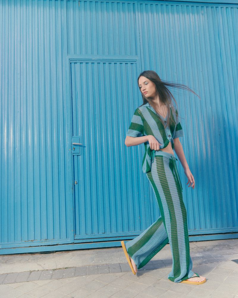 Woman walking in a green and blue striped knit short-sleeve shirt and matching wide-leg trousers past a corrugated blue metal wall.
