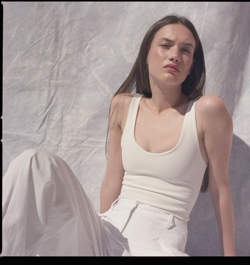 Woman in an off-white scoop-neck tank top and matching wide-leg trousers seated against a textured fabric backdrop.