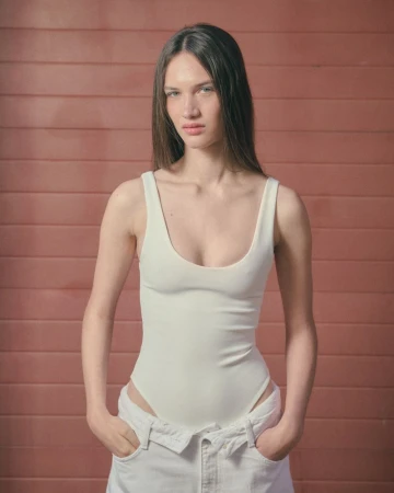A woman with straight dark hair posing in a white scoop-neck bodysuit and matching white trousers against a terracotta slatted wall.