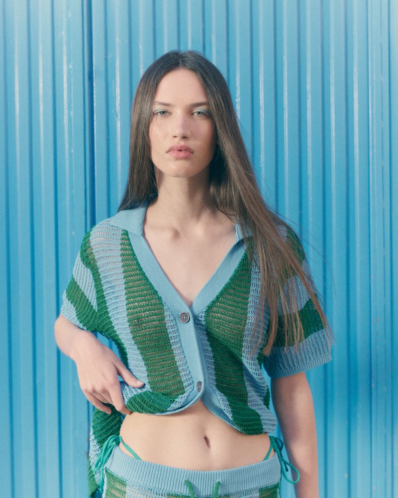 Woman in a green and blue striped knit polo shirt and matching shorts posing against a blue corrugated background.