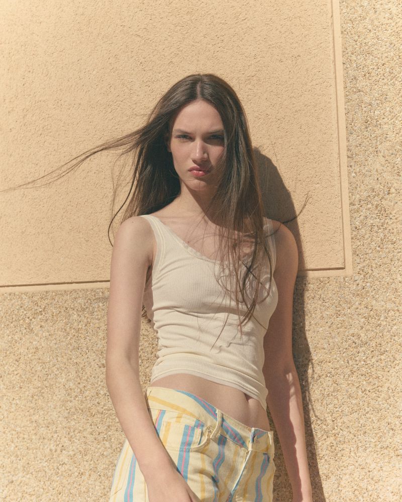 Young woman in an off-white tank top and pastel striped shorts leaning against a sunlit textured wall.