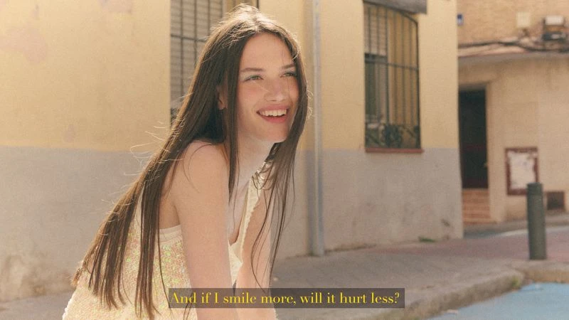 Smiling young woman with long brown hair wearing a pale shimmer top on a sunny city street.