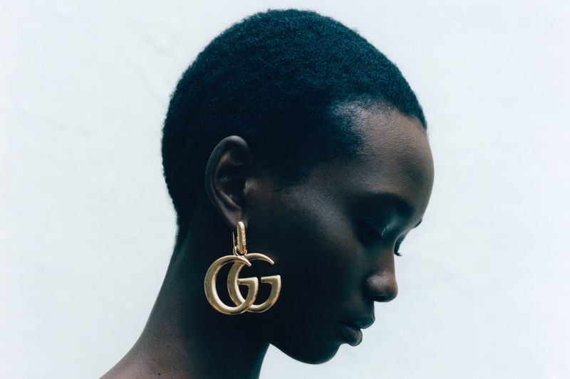 Profile close-up of a Black woman model wearing a large gold Double G drop earring against a bright white background.