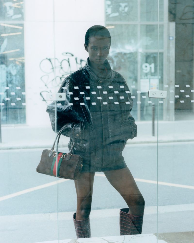 Model Farida Ebene wearing an oversized dark jacket and knee-high boots holding a Gucci handbag behind a glass door.
