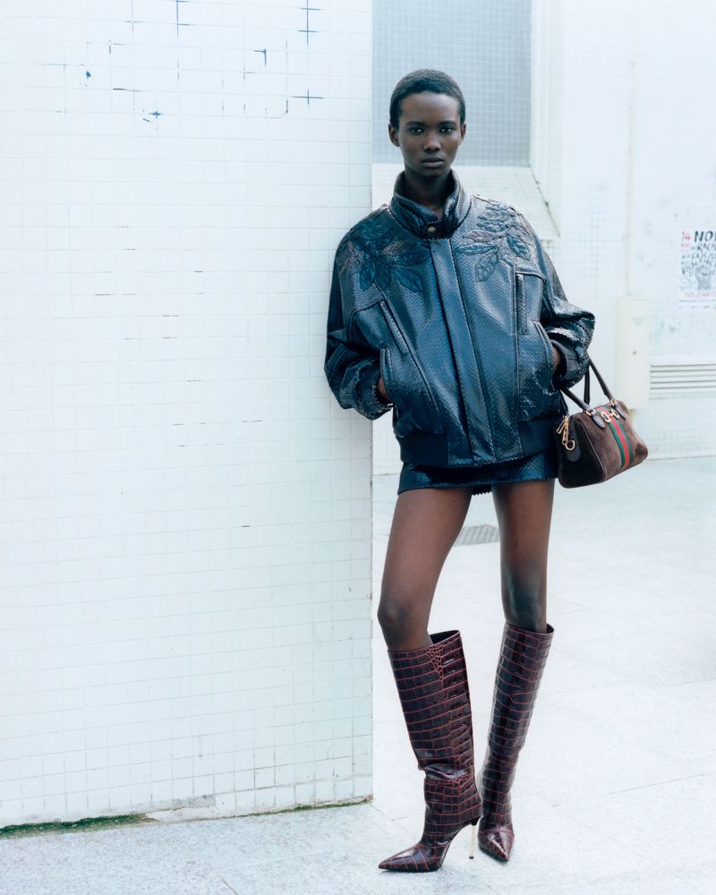Model Farida Ebene poses in an oversized black leather jacket, mini skirt, and knee-high croc boots against a pale tiled wall.