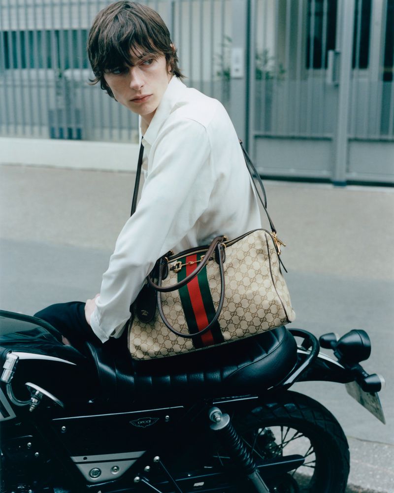 Model sitting on a black motorcycle wearing a white shirt and carrying a monogram canvas duffle bag on a city street.