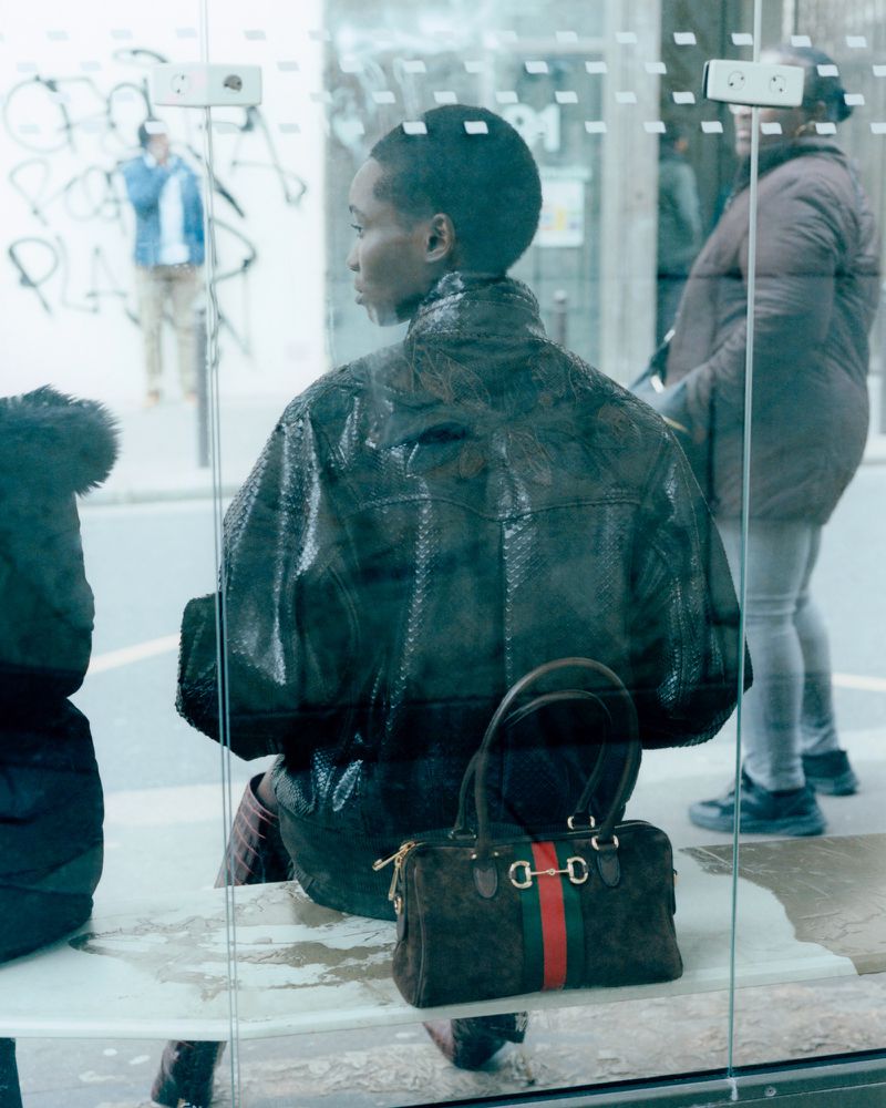Model seen from behind wearing a textured dark jacket sitting at a bus shelter next to a brown handbag with red and green stripes.