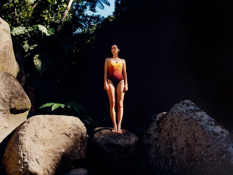 Model Alicia Lindén Guinez standing barefoot on a dark boulder wearing an ombre one-piece swimsuit in natural sunlight.