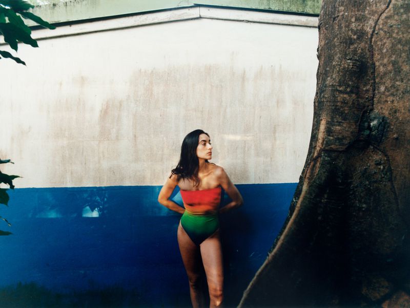 Alicia Lindén Guinez wearing a strapless gradient swimsuit standing beside a large tree trunk against a blue and white wall.