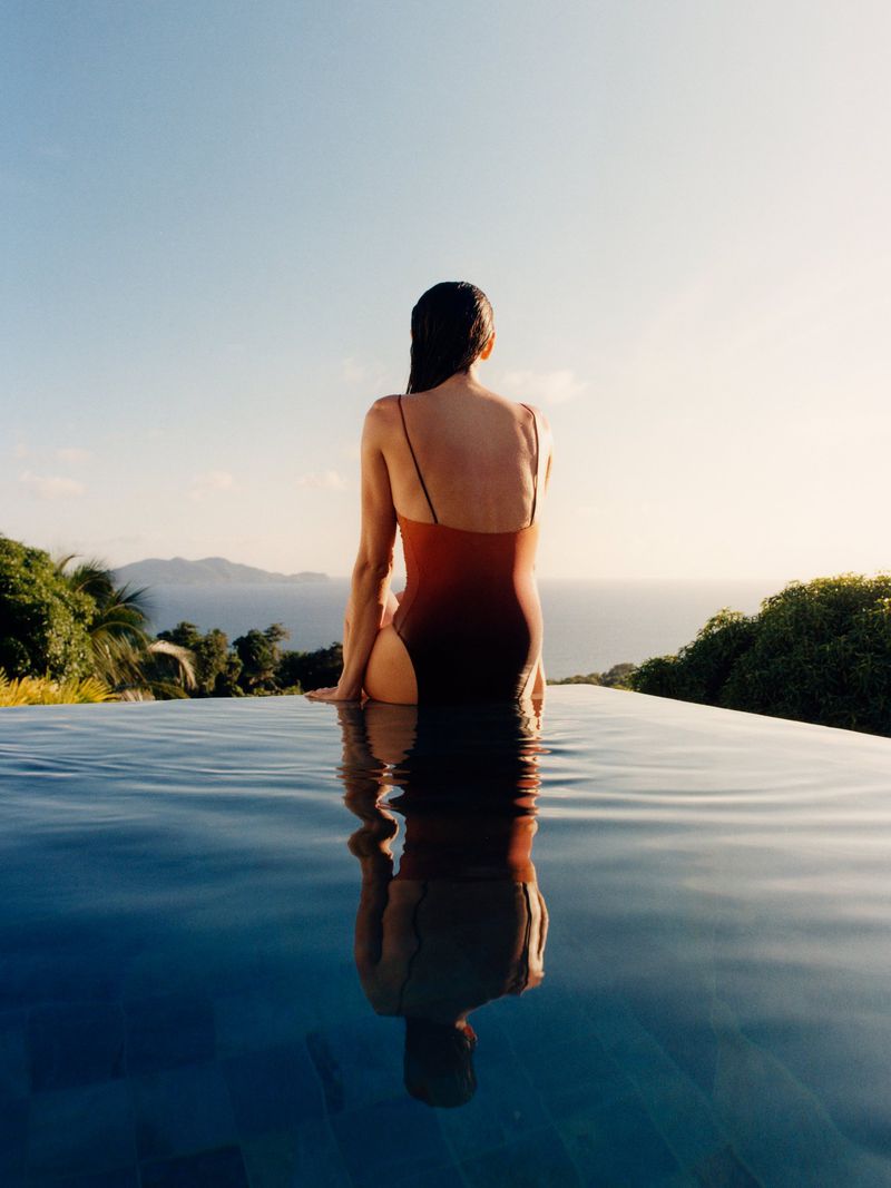 Woman wearing an ombre one-piece swimsuit sitting at the edge of an infinity pool overlooking the ocean.