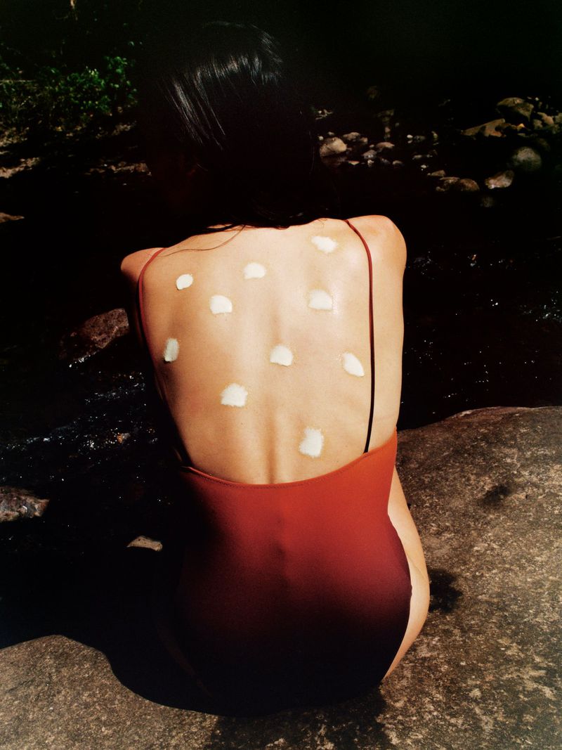 Back view of a woman seated on dark rocks wearing a rust-toned one-piece swimsuit with white lotion dots applied across her back.