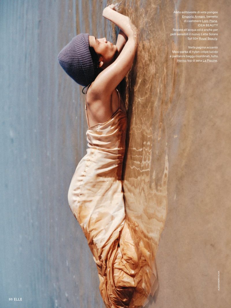 Model wearing a silky slip dress and beanie lying in shallow water on a sandy beach
