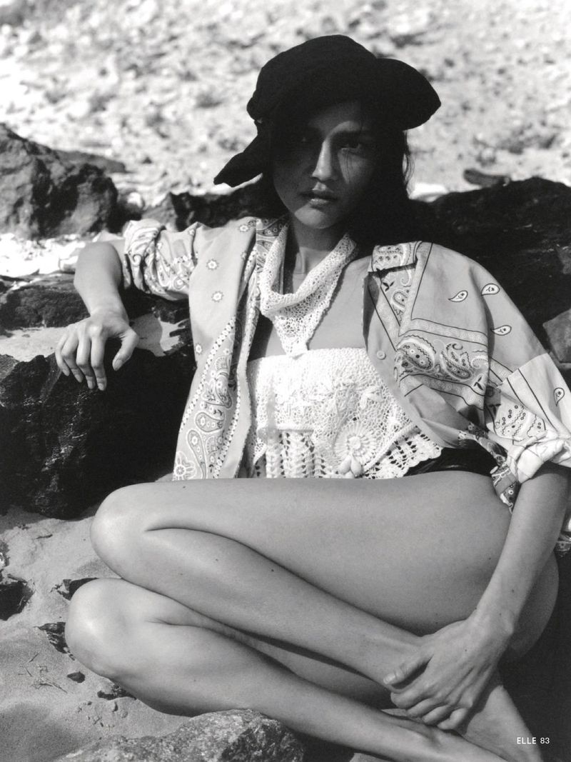 Model wearing a patterned shirt and hat seated among rocky terrain for Elle Italia editorial