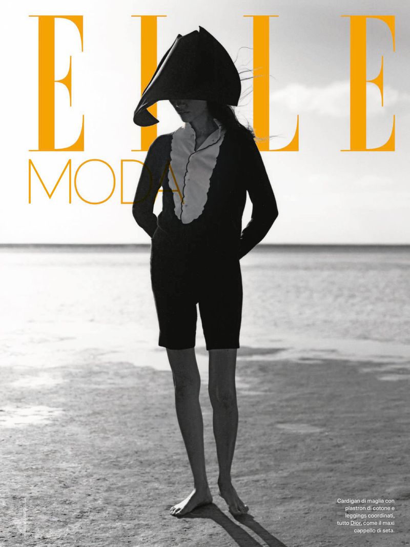 Model Charlotte Carey posing barefoot on a beach in black shorts, a white bib top, and a sculptural black hat for Elle Italia.