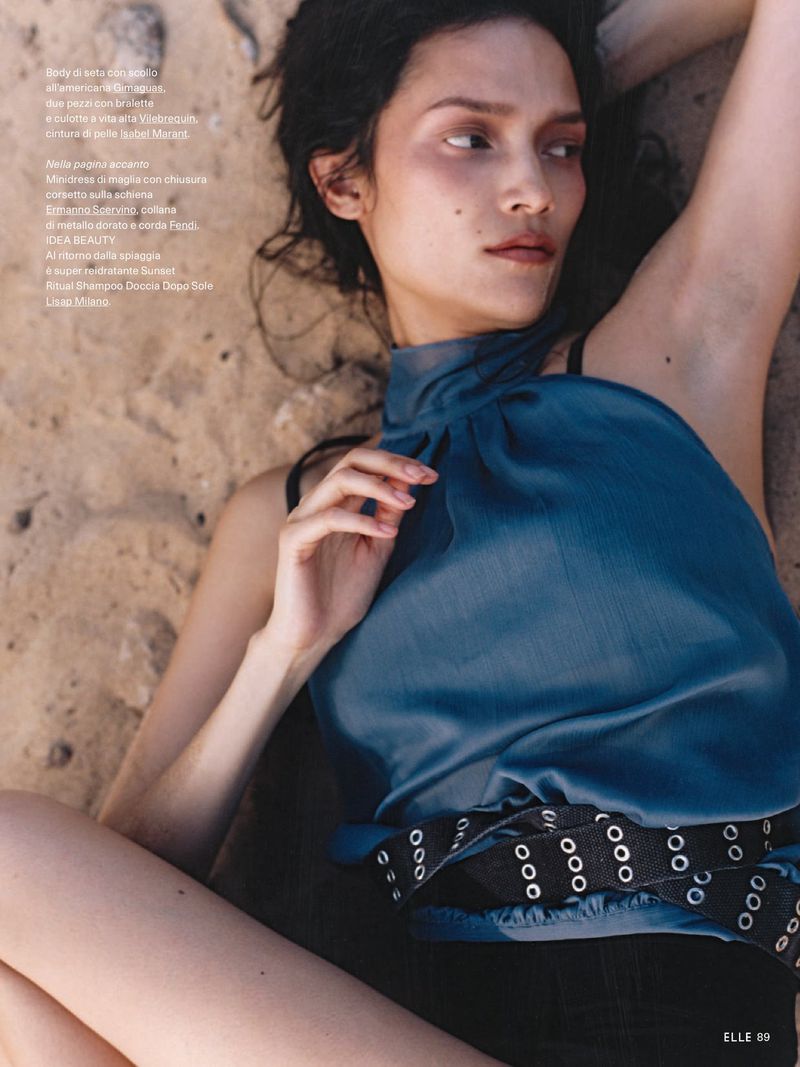 Model Charlotte Carey lying on sandy ground wearing a teal halter top and a black eyelet belt for Elle Italia.