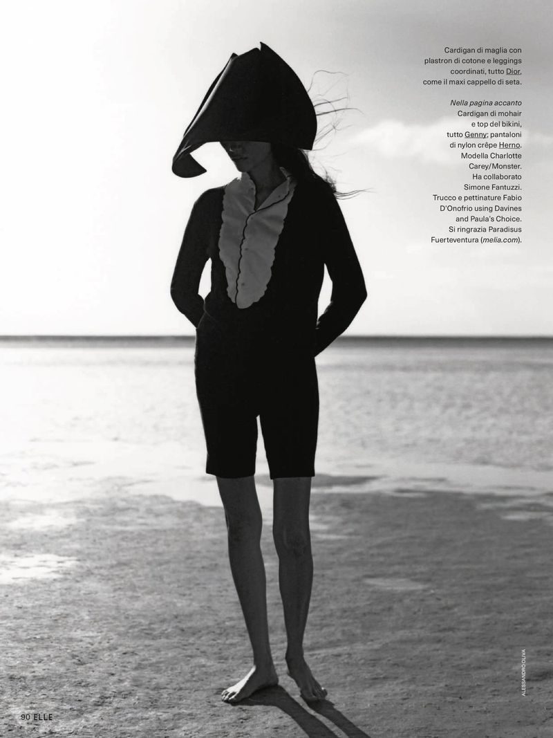 Model Charlotte Carey posing on a sandy beach in a sculptural headpiece, ruffled top, and black shorts for Elle Italia.