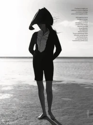 Model Charlotte Carey posing on a sandy beach in a sculptural headpiece, ruffled top, and black shorts for Elle Italia.
