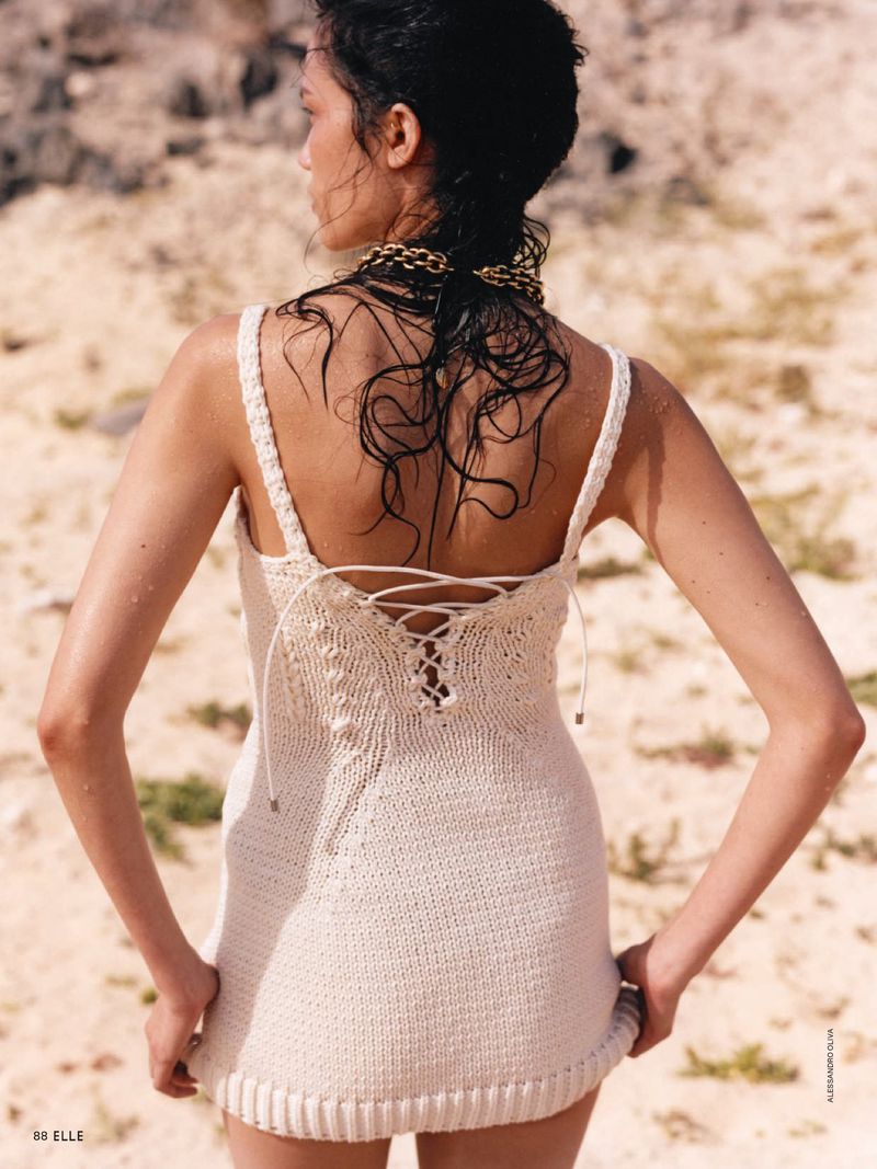 Model Charlotte Carey seen from behind wearing an off-white knitted lace-up mini dress on a sandy outdoor terrain.