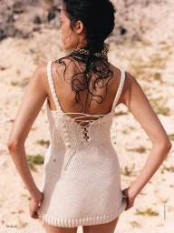 Model Charlotte Carey seen from behind wearing an off-white knitted lace-up mini dress on a sandy outdoor terrain.