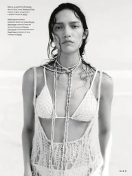 Model with wet hair wearing a string bikini and net knit top accessorized with long layered bead necklaces against an outdoor beach backdrop