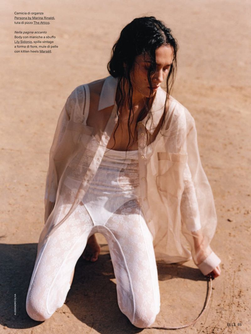 Model Charlotte Carey kneeling on sand in a sheer organza shirt and white lace bodysuit for an Elle Italia editorial.
