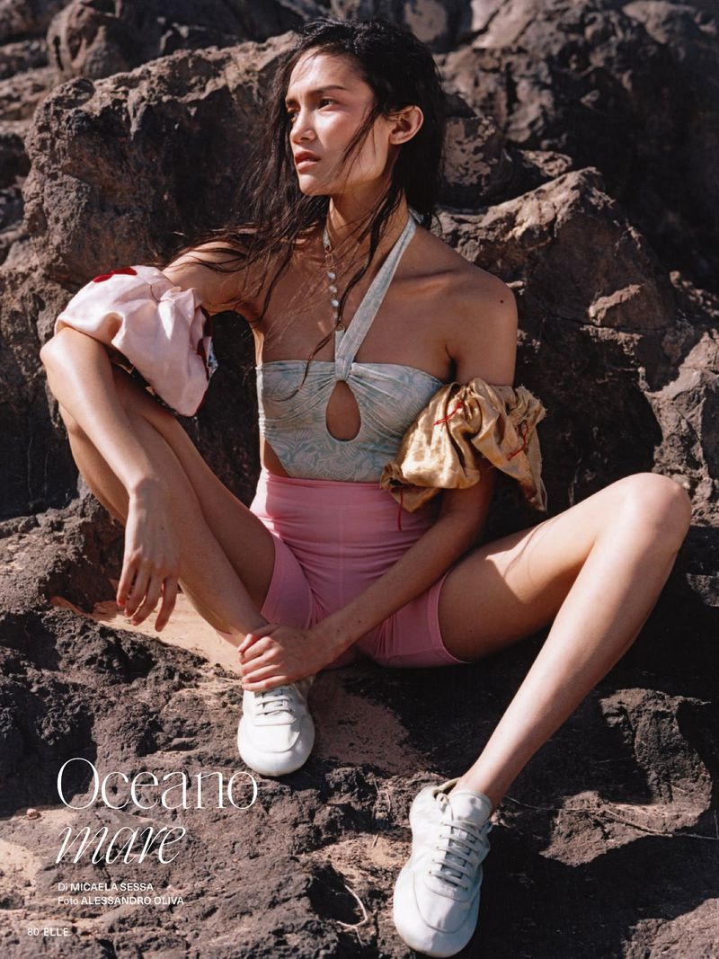 Model Charlotte Carey seated on rugged coastal rocks wearing pink bike shorts, halter top, and white sneakers for Elle Italia.