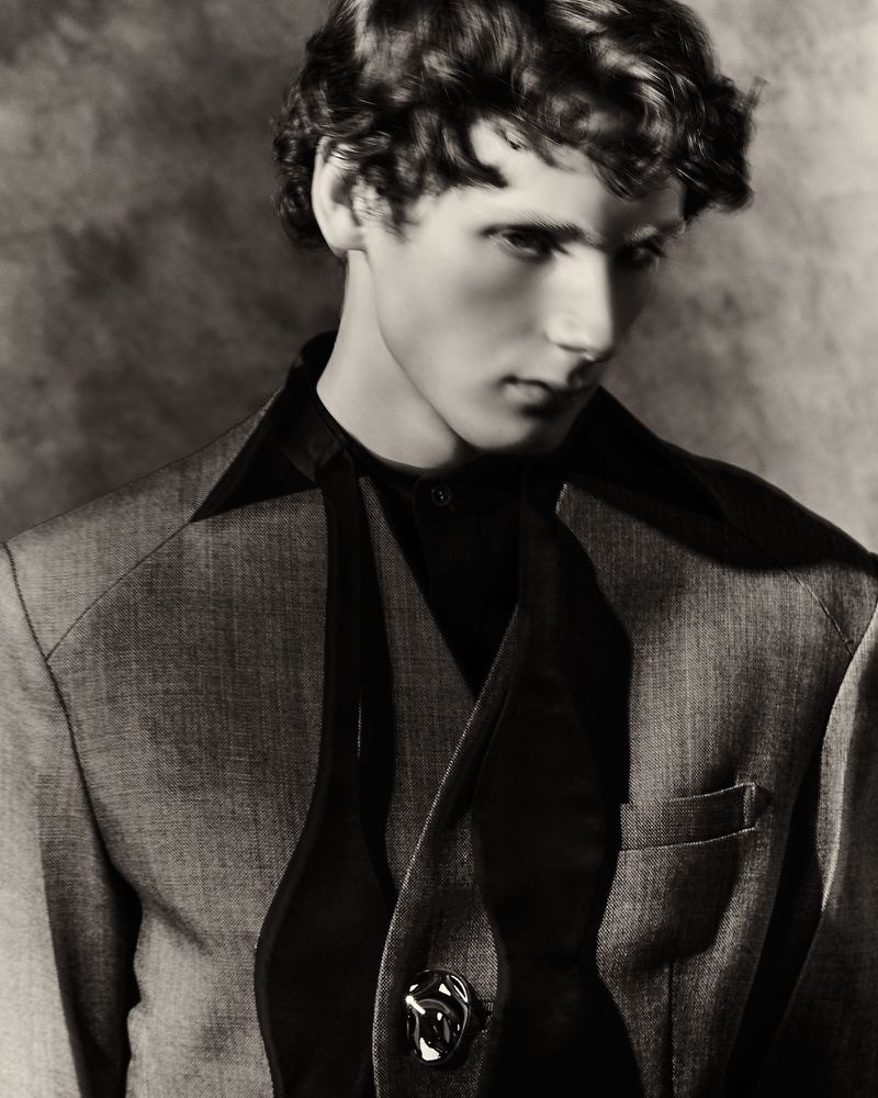 Portrait of a man with wavy hair wearing a tailored grey suit jacket with exaggerated black lapels against a textured backdrop.