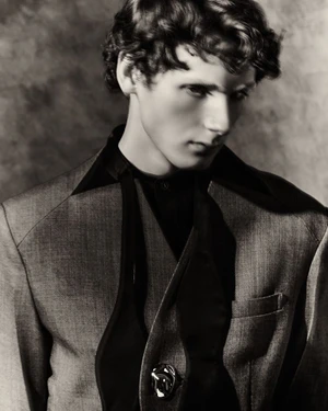 Portrait of a man with wavy hair wearing a tailored grey suit jacket with exaggerated black lapels against a textured backdrop.
