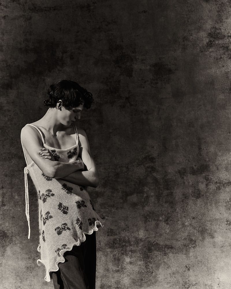 Model with arms crossed wearing a floral knit asymmetrical camisole top against a textured dark backdrop.