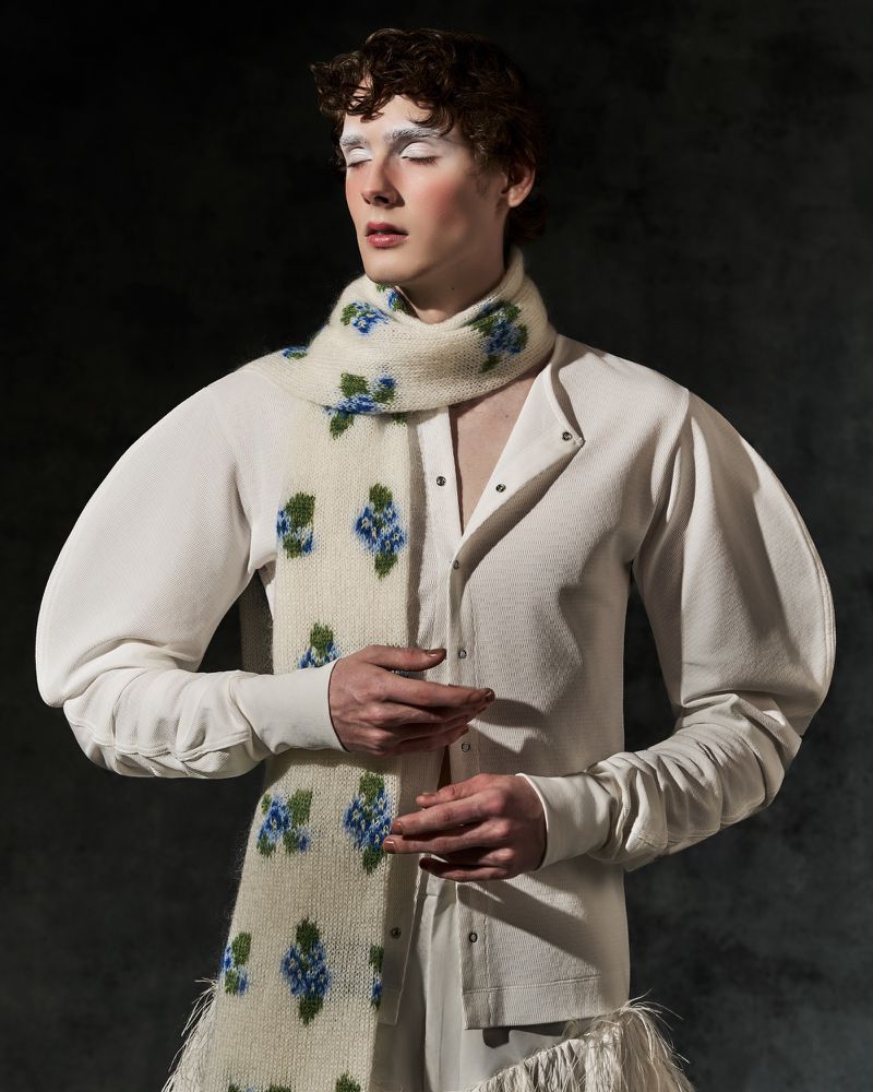 Man with white painted eyebrows wearing a cream puff-sleeve top and floral knit scarf against a textured dark background.