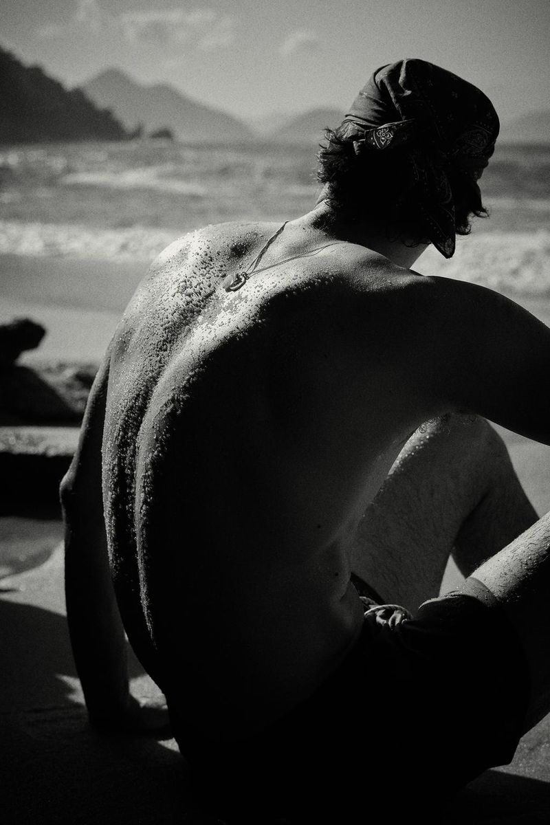 Monochrome shot of model Francesco Ruggiero seated shirtless on a sandy beach looking out toward the ocean waves.