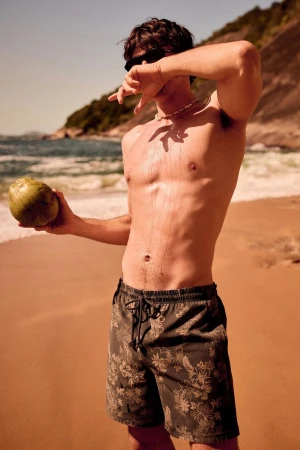 Shirtless man in printed swim shorts holding a green coconut while shielding his eyes on a sunny beach.