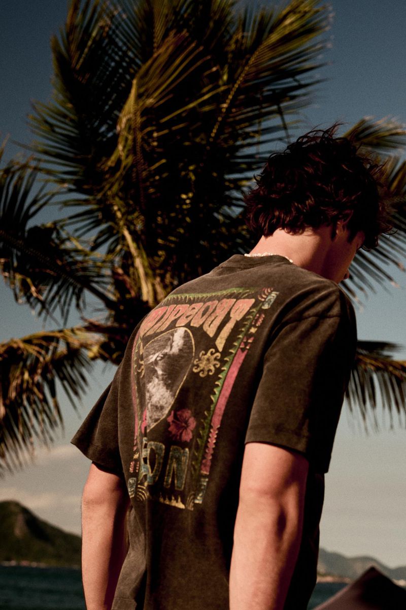 Man from behind wearing a washed dark graphic t-shirt in front of a palm tree and ocean background.