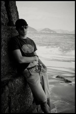 Monochrome photo of a man in sunglasses, t-shirt, and denim shorts leaning against a rock on a beach.